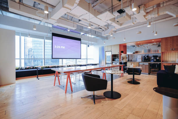 A vibrant and modern co-working space with a mix of comfortable lounge seating and work tables, offering a stylish yet functional environment for collaboration and productivity. The space features a large open area with a combination of wooden furniture, colorful cushions, and indoor plants that create a welcoming atmosphere. The industrial ceiling design with exposed pipes adds a contemporary touch, while the large windows allow ample natural light to flood the room. The blend of traditional and modern elements, such as the wooden partition screens and sleek furniture, contributes to a dynamic and creative workspace.