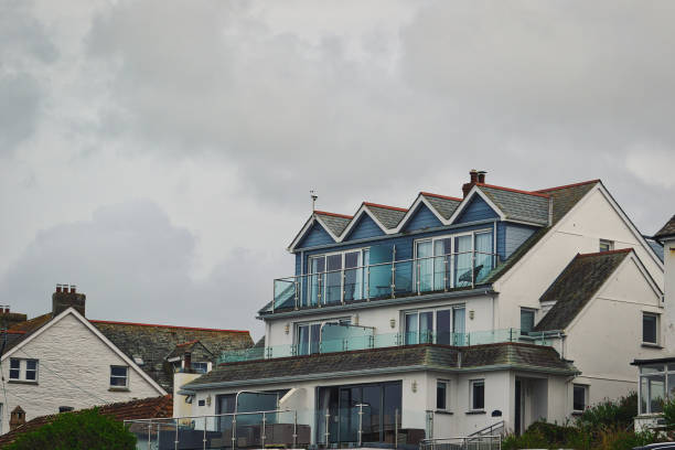 A modern coastal house with multiple balconies and large windows, set against a cloudy sky. The architecture features a unique roof design and is surrounded by other buildings.