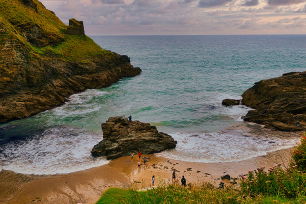 A scenic coastal view featuring a sandy beach surrounded by rocky cliffs. People are enjoying the beach, with gentle waves lapping at the shore. The sky is partly cloudy, adding to the serene atmosphere.