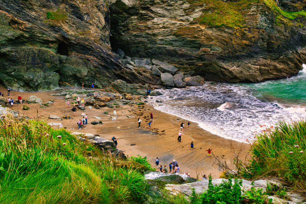 A scenic beach view with people enjoying the shore, surrounded by rocky cliffs and lush greenery. Waves gently lap at the sandy beach, creating a tranquil atmosphere.