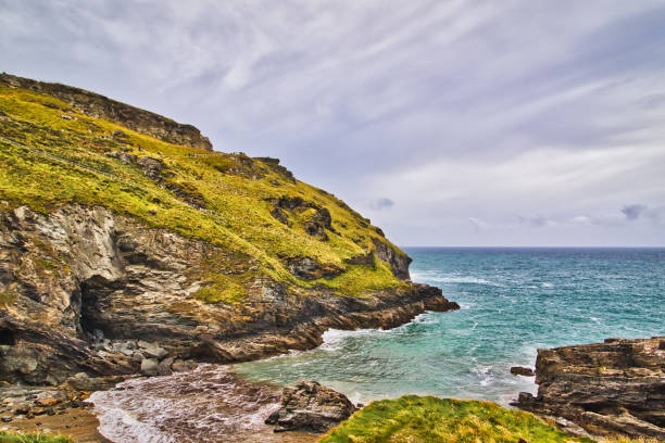 A scenic coastal view featuring rugged cliffs, lush green hills, and a tranquil sea. The sky is partly cloudy, creating a serene atmosphere.