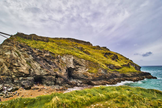 A scenic coastal view featuring a rocky cliff covered in greenery, with a cave entrance visible. The ocean waves crash against the shore, under a cloudy sky.