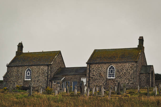 Two stone buildings with mossy roofs, surrounded by a grassy area and gravestones. The sky is overcast, creating a moody atmosphere.