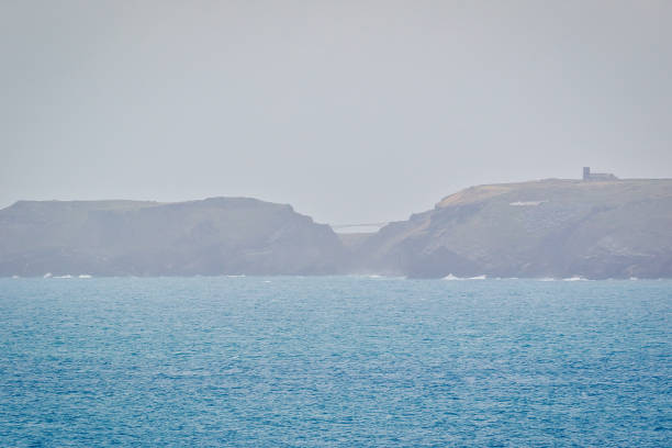 A hazy view of a coastal landscape featuring cliffs and a distant structure on the horizon. The ocean is calm with gentle waves, creating a serene atmosphere.