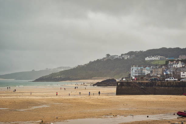 A coastal scene featuring a sandy beach under a cloudy sky. People are walking along the shore and near the water, while a rocky pier is visible in the foreground. Houses are perched on a hill in the background, adding to the scenic view.