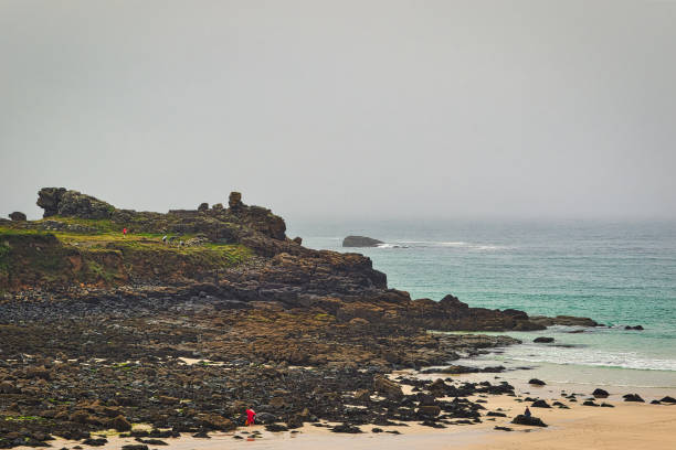 A scenic coastal view featuring rocky shorelines and gentle waves. A person in red is exploring the beach, surrounded by dark rocks and green grass. The sky is overcast, creating a moody atmosphere.