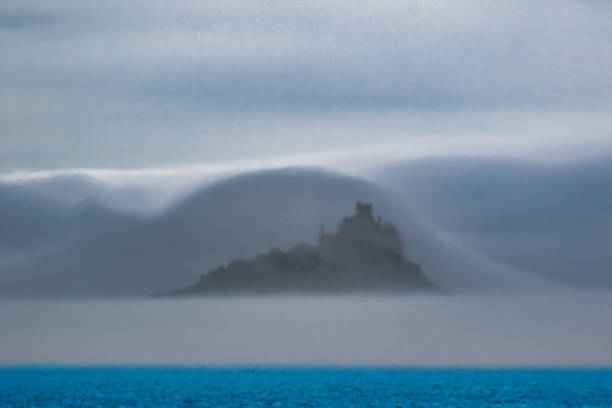 A mystical island shrouded in fog, partially obscured by clouds, with a castle-like structure visible on the island. The scene is tranquil, with a calm blue sea in the foreground and soft gray tones in the background.