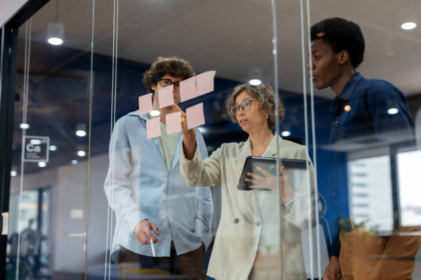 A business development team gathers in a modern office meeting room, brainstorming and discussing their business process improvement strategy. The team, consisting of a diverse group of professionals, uses post-it notes to organize ideas and refine.