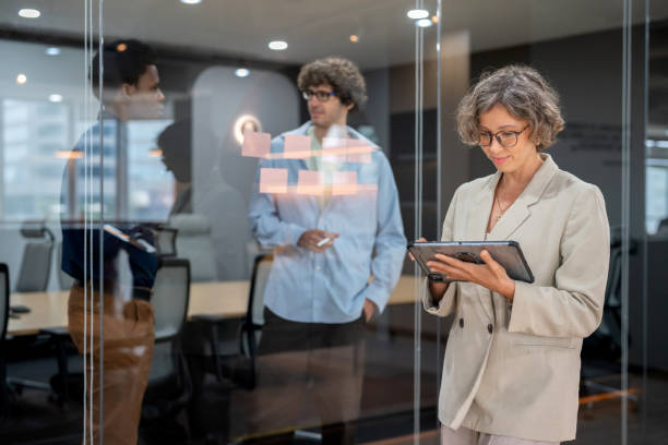 istock Businesswoman walks through a sleek, modern office while reviewing information on digital tablet. She glances through the glass-walled conference room where a group of professionals is standing and engaged in a lively discussion about work. 2173725928