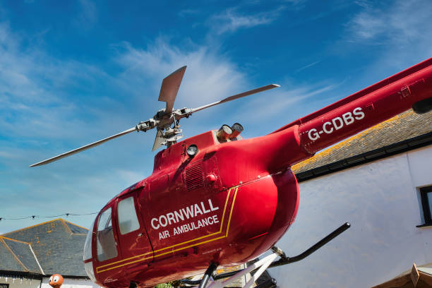 A red helicopter with the words 'Cornwall Air Ambulance' on its side, parked against a clear blue sky. The helicopter's rotor blades are visible, showcasing its design and purpose.