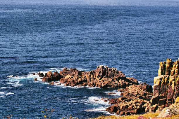 A scenic view of rocky coastline with waves crashing against the rocks, surrounded by deep blue ocean water. The rocks display various colors and textures, creating a natural landscape.