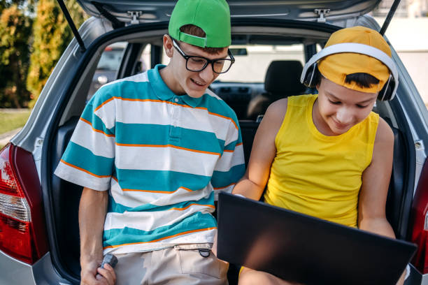 Teenagers sitting in car trunk using laptop before leaving on sport training