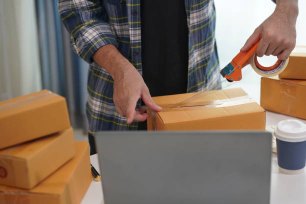istock Small business owner starting out using a smartphone and writing addresses on cardboard boxes and packing parcels for home delivery. Small business delivering parcels to customers. SME entrepreneur. 2173825100
