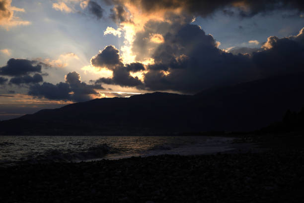 A dramatic sunset illuminates dark clouds over a rocky beach. The light rays break through the clouds, reflecting on the sea and highlighting the distant horizon.
