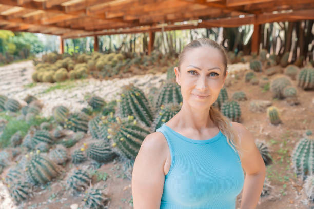 Smiling woman in a blue tank top standing in a cactus garden under a wooden structure, surrounded by various cacti.