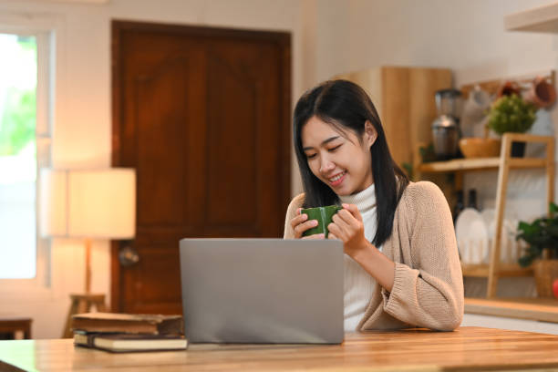 istock Smiling young asian woman sitting at kitchen table and using her laptop 2173863672