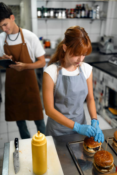 Teenage girl making burgers int he fast food restaurant kitchen, while her coworker taking orders Caucasian redhead female teenager making burgers at the fast food restaurant, while her male teenage coworker taking inventory or orders on a digital tablet girls getting ready to go out stock pictures, royalty-free photos & images