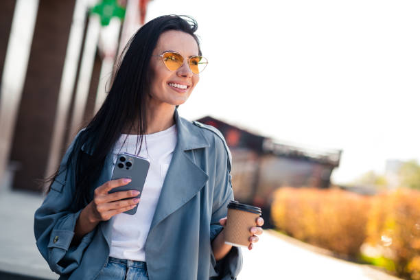 istock Photo of cheerful pretty lady dressed grey jacket texting modern device walking drinking tea enjoying sunshine outdoors town street 2173896976