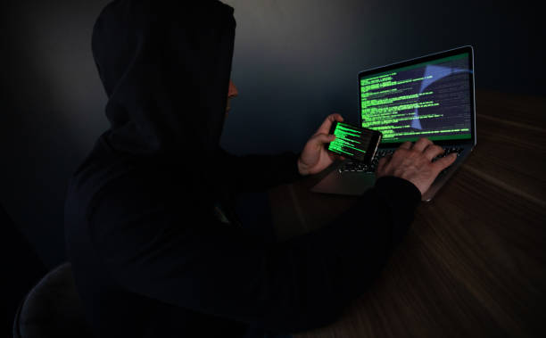 istock A hooded programmer person sits at a desk using a laptop and smartphone with green code displayed, suggesting hacking or cybersecurity activity. The dark setting adds a sense of mystery and secrecy. 2173912363