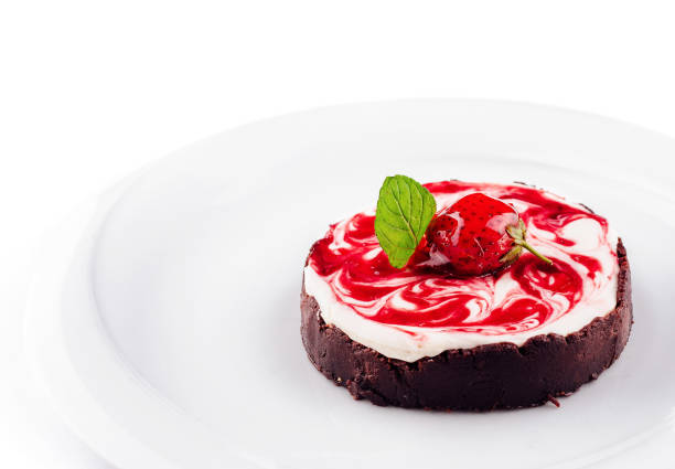 Delicious cheesecake with strawberry sauce is sitting on a white plate, topped with a strawberry and mint leaf