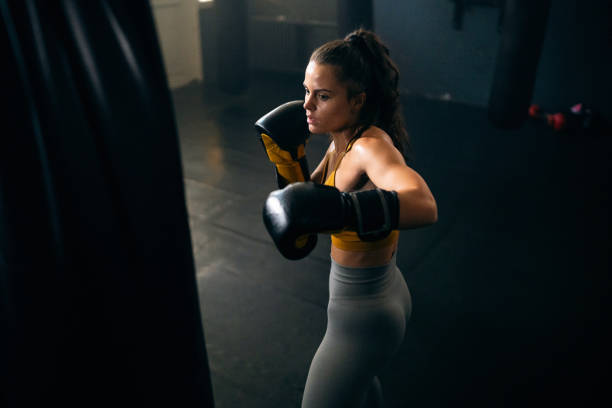 istock Young female athlete, with boxing gloves, punching the punching bag, while practicing kickbox in the gym 2173926777