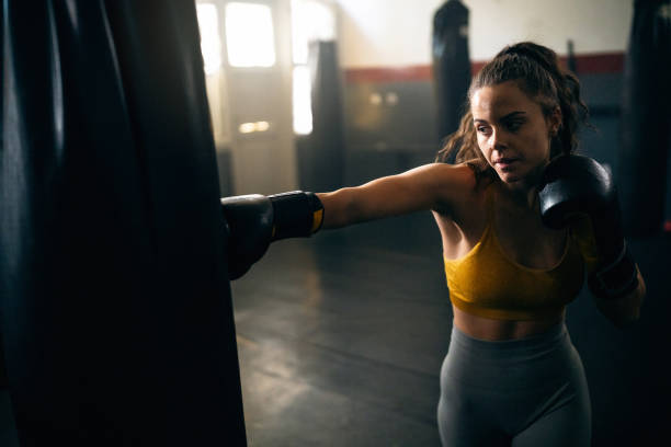 istock Caucasian female athlete, with boxing gloves, punching the punching bag, while practicing kickbox in the gym 2173929114