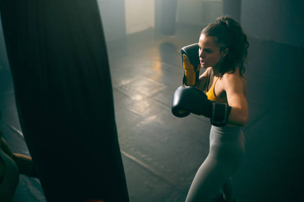 istock Young female athlete, with boxing gloves, punching the punching bag, while practicing kickbox in the gym 2173929510