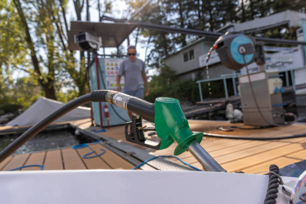 Close-up of a fuel nozzle on a boat at a lakeside station, with a man walking towards it. Trees and marina visible in the background.