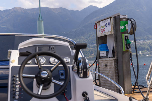 Steering wheel and controls of a boat docked at a marina, with a fuel station in the background against a backdrop of mountains and calm water.