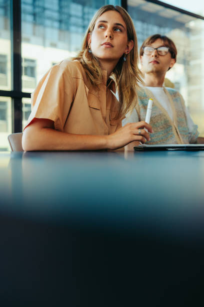 Female caucasian student concentrating in class while holding a marker, focused on learning in a modern educational setting with peers