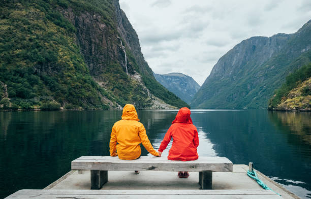 Happy couple dressed in bright windproof jackets sitting and hugging on bench on the Norwegian NÃ¦rÃ¸yfjord bank in Gudvangen, Norway. Traveling, beauty in Nature and relations concept.