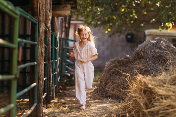 A happy young girl in a white dress joyfully runs through a rustic farm barn, surrounded by hay and natural sunlight.