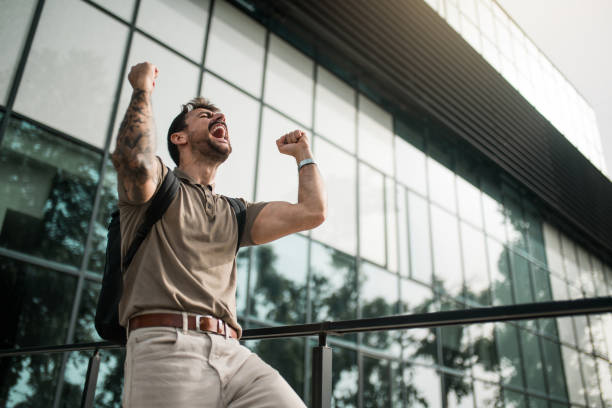 Ecstatic young man celebrating success while standing in front of an office building. Low angle view of ecstatic young male entrepreneur celebrating success with hands up while standing in front of an office building. arm tattoo ideas for men pictures stock pictures, royalty-free photos & images