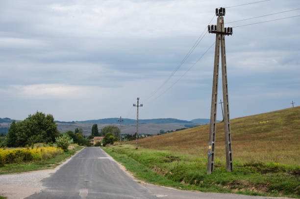 A quiet rural road lined with power poles and wires, stretching through the countryside with grassy fields on either side. Rolling hills and distant trees complete the tranquil scene under a cloudy sky.