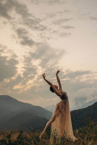 This evocative photograph portrays a dancer elegantly stretching with arms reaching towards the dramatic sky, set against a backdrop of majestic mountains and a dusky evening sky. The dancer, clad in a flowing, semi-transparent gown, appears to be in perfect harmony with the natural surroundings. The scene is beautifully illuminated by the soft, diffused light of the setting sun, casting a warm glow over the landscape and the dancer. The image captures the essence of grace, freedom, and the profound connection between human movement and the timeless beauty of nature.
