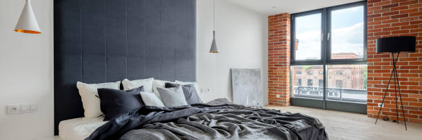 Panorama of spacious and elegant bedroom with industrial style wall with exposed red brick, big comfortable bed with upholstered wall and big window