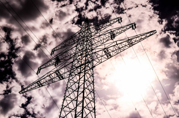A tall power tower is silhouetted against a cloudy sky. The sky is overcast and the sun is barely visible