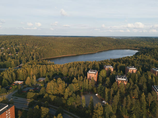 An aerial view of a residential area nestled within a vast forest landscape, featuring apartment buildings surrounded by trees and a serene lake in the background. The image showcases the harmonious coexistence of urban development and natural environment under a partly cloudy sky.