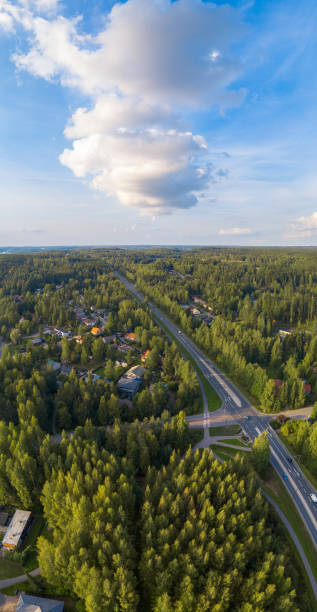 An aerial photograph showcasing a lush green forest landscape intersected by a winding highway. Small clusters of houses are visible among the trees, indicating a rural or suburban community. The image is topped with a bright blue sky adorned with fluffy white clouds, creating a picturesque scene of nature and human development coexisting.