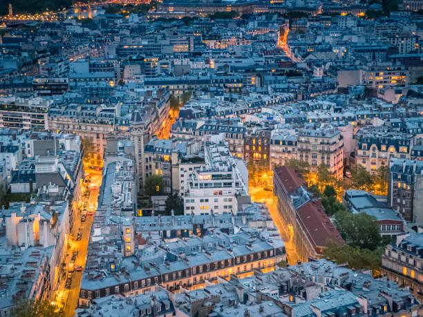 An aerial view of Paris at night, showcasing the city's dense urban landscape illuminated by streetlights and building lights. The image captures the iconic Parisian architecture, with its uniform Haussmann-style buildings, intricate street patterns, and the warm glow of the city after dark
