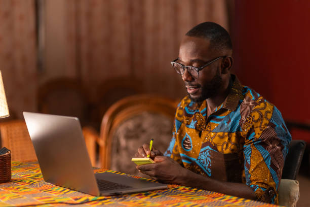 istock An African man in a colorful shirt sits at a table with a laptop open, taking notes while looking focused. 2174019900