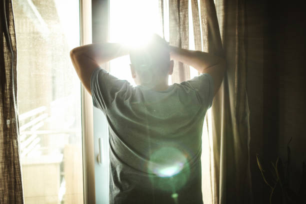 Rear view of adult man looking through window in bedroom
