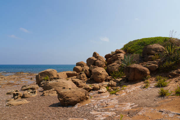 A scenic Mediterranean coastal landscape featuring rugged rocks, coastal vegetation, and the blue sea under a clear sky, highlighting the natural beauty of the shoreline