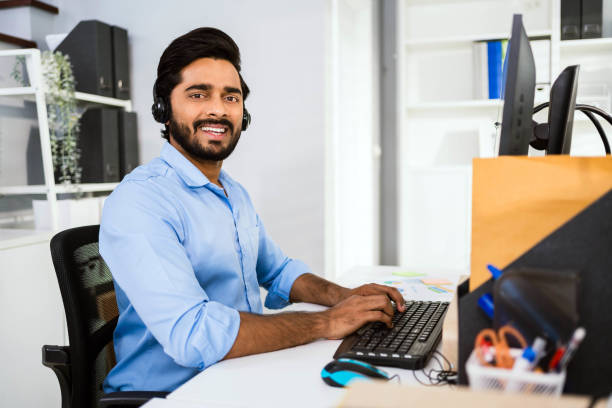 istock A smiling Indian man with headphones works at a desk, using a computer in a bright office environment. 2174076313