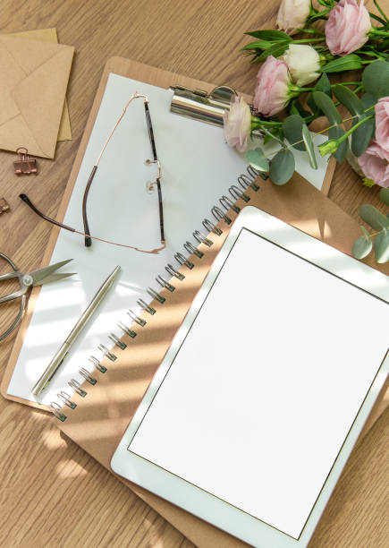 Stationery workspace with a blank tablet, notebook, and flowers on a wooden table, perfect for creative planning in natural light