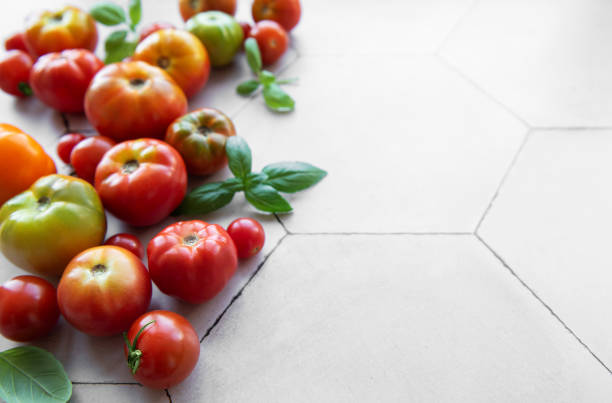 A variety of heirloom tomatoes, including red, yellow, and green, are arranged casually on a light-colored hexagonal tiled surface alongside vibrant basil leaves.