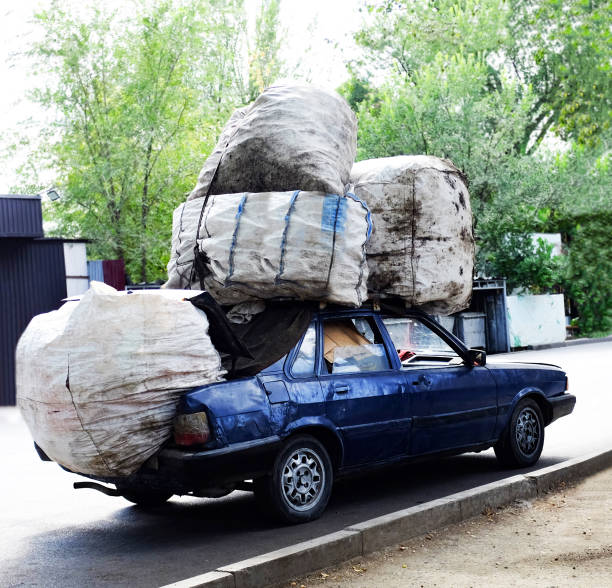 An old blue car is standing on the road, huge bags are installed on its roof and trunk, it is bent from gravity, the windshield is cracked.