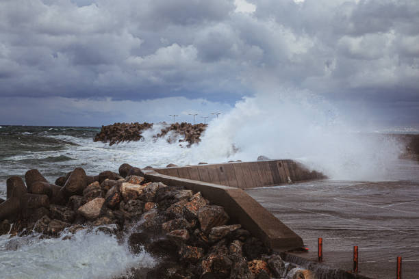 A powerful wave crashes over a harbor breakwater, sending a surge of water into a flooded parking lot nearby. The scene, set during a storm in Rethimno, Crete, captures the dramatic interaction between the turbulent sea and urban infrastructure. Dark, ominous clouds loom overhead, while the churning ocean contrasts with the stillness of the rocks and parking bollards, highlighting the impact of the storm on the coastal landscape.