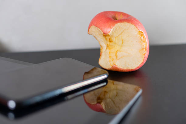 A bitten red apple rests on a black surface next to two smart phones. The apple shows its interior with a shiny skin, while the smart phones reflect the apple and the surface.