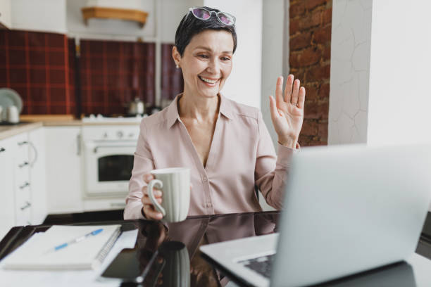 istock Working from home. Mature elegant smiling female executive manager working from home, video chatting with her colleagues, greeting and waving hand, sitting at kitchen table with coffee cup 2174089449